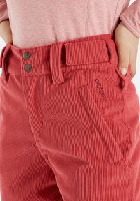 Rote Cordhose mit zwei großen Knöpfen, einem Gürtelschlaufen und einer seitlichen Tasche. Der Stoff hat eine senkrechte Rippstruktur. Gesticktes Logo sichtbar.