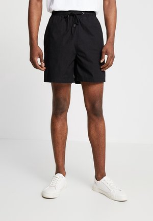 Mann trägt schwarze Shorts mit Kordelzug, weiße Sneakers und ein weißes Hemd, steht vor einem einfarbigen hellen Hintergrund.