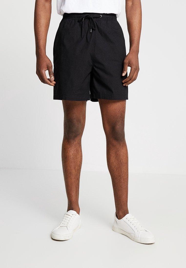 Homme portant un short noir à cordon, des baskets blanches et une chemise blanche, debout devant un fond clair uni.