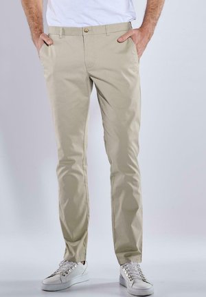 Beige Chinos mit schlanker Passform, ausgestattet mit Vorder-taschen, einem einzigen Knopfverschluss und glattem, leichtem Baumwollstoff. Kombiniert mit weißen Sneakers.
