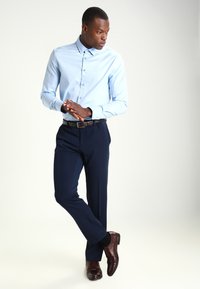 Camisa de botones azul claro, pantalones navy y zapatos de cuero marrón. La camisa tiene un cuello; los pantalones son ajustados y tienen una textura suave.