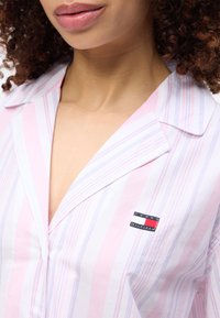 Γυναίκα που φοράει ριγέ ροζ και λευκό πουκάμισο με γιακά, με ένα μικρό ορθογώνιο λογότυπο Tommy Hilfiger στην αριστερή πλευρά.