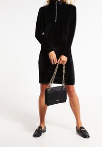 Schwarzes Samtkleid mit hohem Kragen und Reißverschlussdetail, kombiniert mit einer schwarzen gesteppte Handtasche mit Kettenriemen und flachen Schuhen.