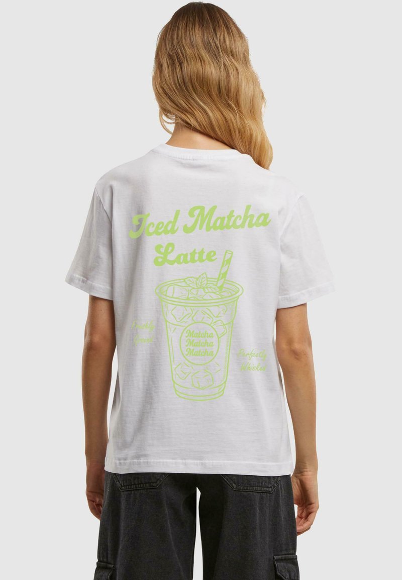 Balta medvilninė marškinėliai su neoninės žalios spalvos grafika, vaizduojančia šaltą matcha latte, ir tekstu. Trumpomis rankovėmis ir laisvu kirpimu.
