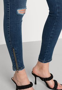 Jeans skinny em azul escuro com joelho desgastado. Apresenta um fecho dourado no tornozelo, combinado com sandálias de salto pretas.