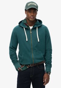 Sudadera con cremallera en color verde azulado con cordones blancos, llevada sobre una camisa de cuadros. Jeans oscuros y una gorra verde con el logo "Superdry", que incluye un reloj.
