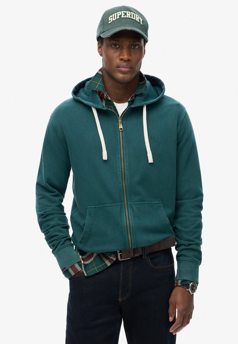 Sudadera con cremallera en color verde azulado con cordones blancos, llevada sobre una camisa de cuadros. Jeans oscuros y una gorra verde con el logo "Superdry", que incluye un reloj.