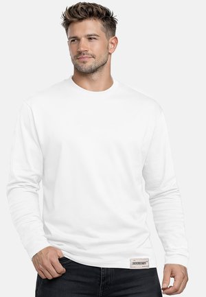 Mann mit kurz gestyltem Haar, trägt ein schlichtes weißes Langarmshirt und schwarze Hose, steht vor einem hellen Hintergrund.