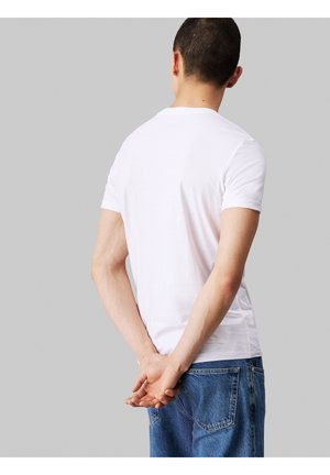 Homme aux cheveux courts portant un t-shirt blanc et un jean bleu, debout les mains croisées dans le dos, de dos sur un fond gris uni.