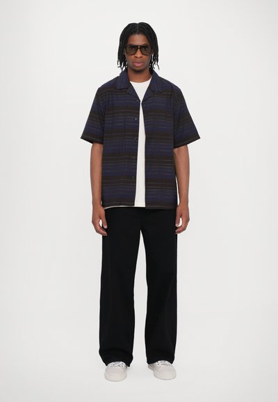 Chemise à manches courtes de couleur bleu foncé avec des rayures horizontales, associée à un pantalon large noir. Le modèle porte des baskets blanches et des lunettes de soleil.