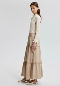 Lange beige jurk met aansluitende corsage, gelaagde rok, decoratieve afwerking en lange mouwen. Draagt met beige platte schoenen. Glad textuur.