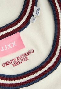 Hvid stofkrave med røde, hvide og blå striber, pink JJXX mærke, størrelse S label og "Made in Bangladesh" trykt mærkat.