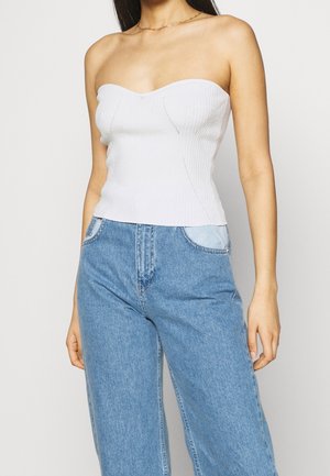 Kobieta ubrana w biały, bezramienny prążkowany top i wysokie niebieskie dżinsy z jasnoniebieskimi detalami kieszeni, stojąca na jednolitym tle.