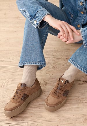 Person wearing light brown sneakers with dark brown details, beige socks, and blue denim jeans seated on a light wooden floor.