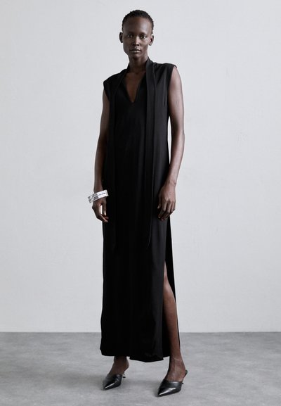 Lungo vestito nero con profondo scollo a V e design senza maniche, caratterizzato da fessure laterali. Accompagnato da braccialetti a manica trasparente e scarpe nere a punta.