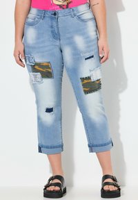 Vaqueros capri de mezclilla en azul claro con parches desgastados, bordes deshilachados y un dobladillo enrollado. Las texturas combinadas incluyen piezas de tejido de camuflaje y liso.