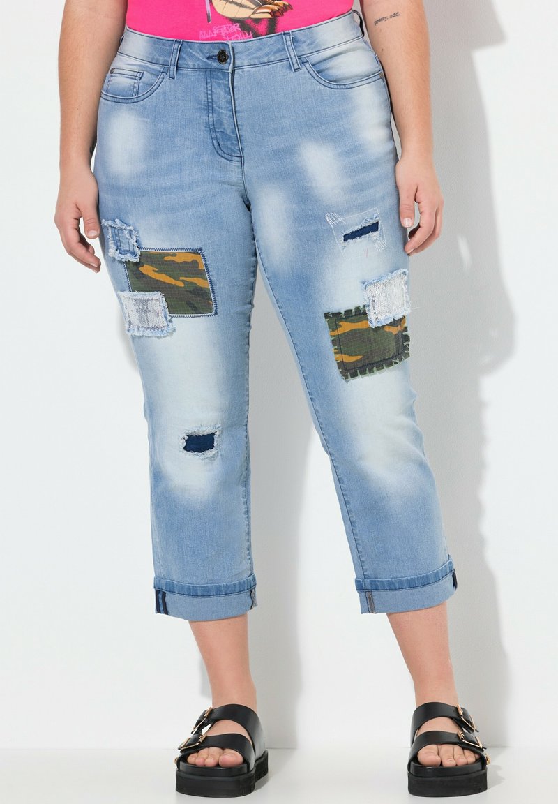Vaqueros capri de mezclilla en azul claro con parches desgastados, bordes deshilachados y un dobladillo enrollado. Las texturas combinadas incluyen piezas de tejido de camuflaje y liso.