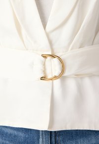 White blouse featuring a draped front, a wide sash belt with a gold circular buckle, and a soft, smooth texture. Paired with denim.