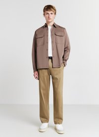 Braunes Hemd mit Knopfleiste und zwei Brusttaschen, weißes T-Shirt darunter, khakifarbene Hose und weiße Sneakers mit beigen Akzenten.