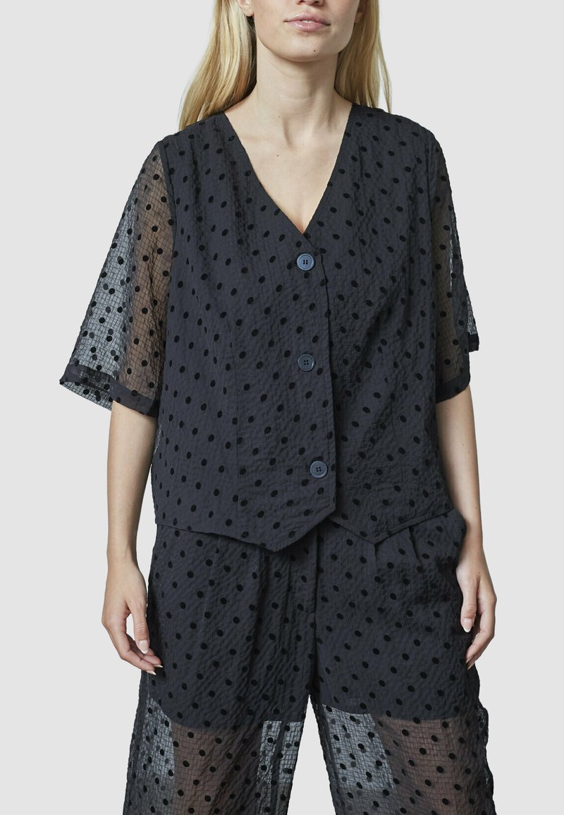 Kvinne iført gennemsigtig sort bluse med polka prikker og knapper samt matchende løstsiddende shorts foran en ensfarvet baggrund.