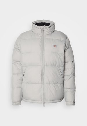 Chaqueta acolchada gris con cuello alto, cierre de cremallera y puños elásticos. Tiene un diseño acolchado y un pequeño parche del logo en el pecho.