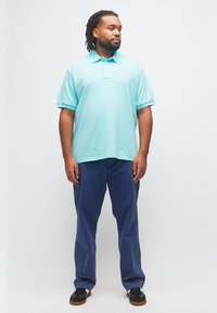 Άνδρας με πλεξούδες, φορώντας ανοιχτό μπλε πόλο μπλούζα, σκούρο μπλε παντελόνι και μαύρα αθλητικά, στέκεται μπροστά σε απλό λευκό φόντο.