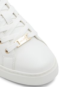 White sneaker made of faux leather, featuring flat laces, a gold-tone logo plate, and textured rubber sole with a round toe.