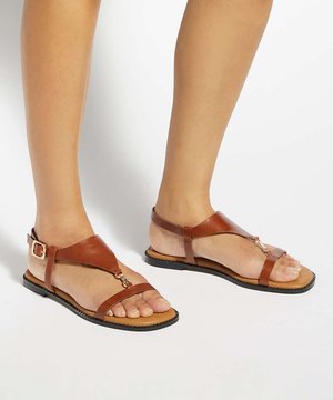 Pieds portant des sandales plates en cuir marron avec des lanières à la cheville et une boucle dorée sur un fond blanc uni.