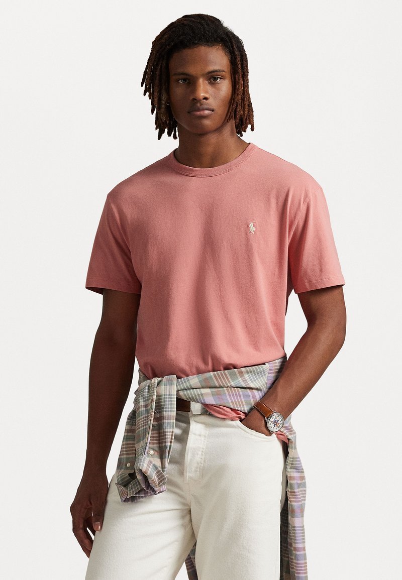 Roze katoenen T-shirt met een ronde hals, voorzien van een klein logo op de borst. Witte broek en een geruite blouse om de taille gebonden.