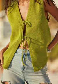 Kobieta w bezrękawnikowej, limonkowej dzianinowej bluzce związanej z przodu dwoma kokardkami oraz w jasnoniebieskich, wysokozakrętnych szortach dżinsowych z przetarciami.