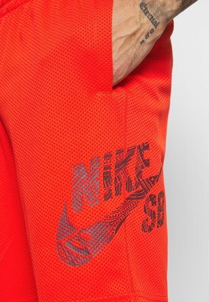 Lyse orange netshorts med et sort mønstret Nike Sports-logo på låret og en hånd, der hviler i lommen.