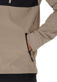 Beige Outdoor-Jacke mit schwarzem Oberteil, ausgestattet mit einer Reißverschlusstasche an der Seite und elastischen Bündchen. Das strukturierte Material sorgt für Langlebigkeit.