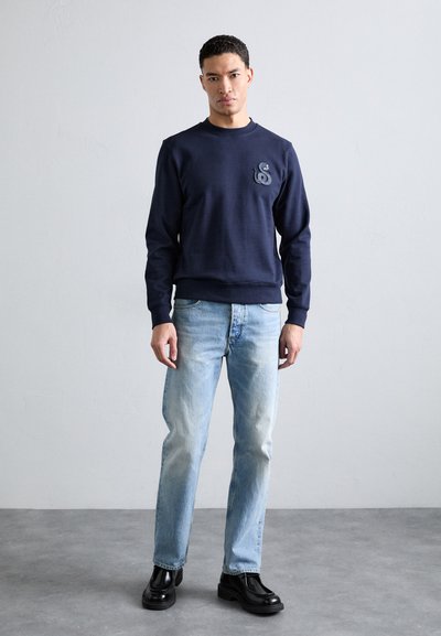 Felpa blu navy con scollatura rotonda, caratterizzata da un logo testurizzato sul petto. Indossata con jeans azzurri e scarpe nere.