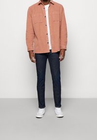 Rosa kordflodsskjorta med knappstängning framtill och två bröstfickor, kombinerad med mörka jeans och vita sneakers. Enkel design, avslappnad passform.