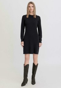Abito nero a maglia fino al ginocchio con maniche lunghe, scollo a girocollo e silhouette aderente. Abbinato a stivali alti in suede scuro.