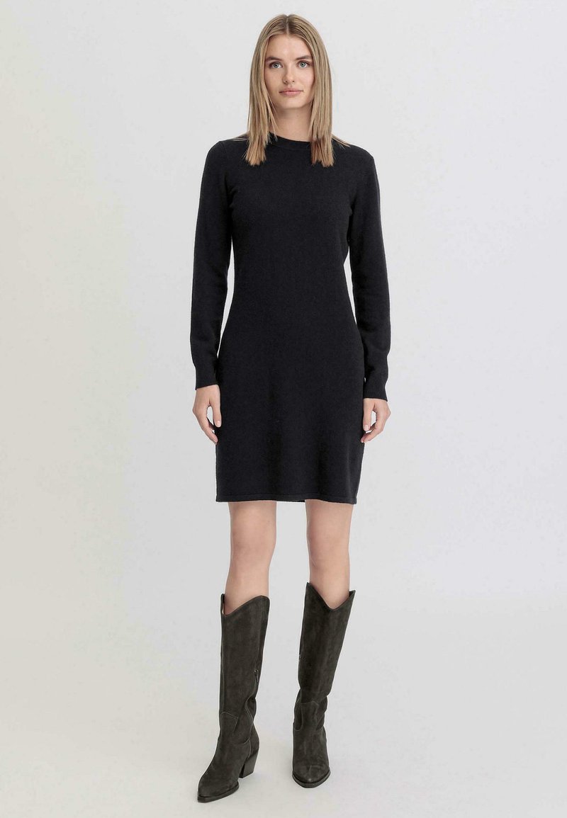 Abito nero a maglia fino al ginocchio con maniche lunghe, scollo a girocollo e silhouette aderente. Abbinato a stivali alti in suede scuro.