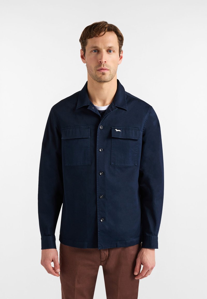 Chemise bleu marine avec col, manches longues et deux poches avant, ornée d'un petit logo blanc et d'une fermeture par boutons. Texture de tissu lisse.