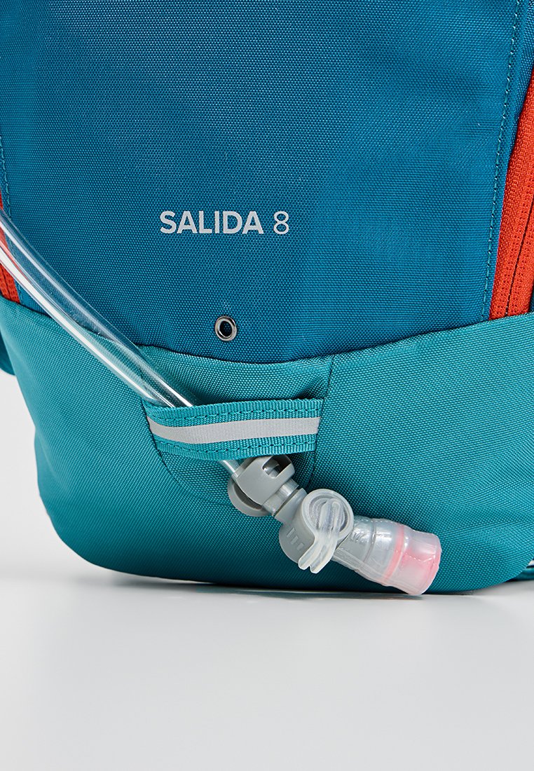 Blå och aqua ryggsäck med röda detaljer, utrustad med en vätskeslang och ventil, stor huvudfack och "SALIDA 8" text på framsidan.