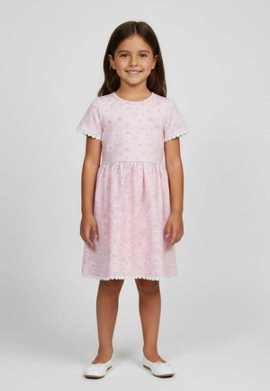 Uśmiechnięta dziewczyna z długimi brązowymi włosami, ubrana w różową sukienkę w kwiaty z krótkim rękawem i białe baleriny, stoi na jednolitym tle.