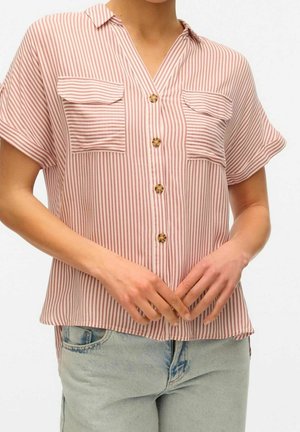 Kobieta w koszuli z krótkim rękawem zapinanej na guziki, z czerwono-białymi pionowymi paskami i dwoma kieszeniami na piersiach, w połączeniu z jasnoniebieskimi dżinsami.