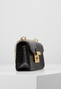 Handväska i krokodilmönster i svart och grått, med ett lås och kedjerem i guldton; har en strukturerad form och glänsande textur.