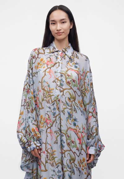 Jeune femme aux longs cheveux foncés portant une robe ample, boutonnée, bleu clair avec des motifs floraux et de vigne colorés.
