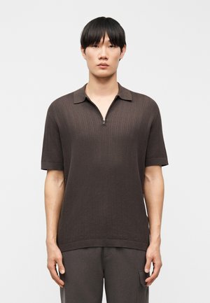 Junger Mann trägt ein dunkelbraunes kurzärmeliges Strick-Poloshirt mit halbem Reißverschluss, steht vor einem schlichten hellen Hintergrund.