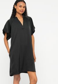 Vestido preto de mangas curtas com decote em V aberto, corte descontraído, bolsos laterais e tecido suave. Textura lisa com uma silhueta ligeiramente solta.