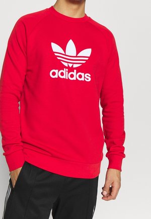 Czerwona bluza z długim rękawem, okrągłym dekoltem, z dużym białym logo Adidas i graficznymi paskami na klatce piersiowej.
