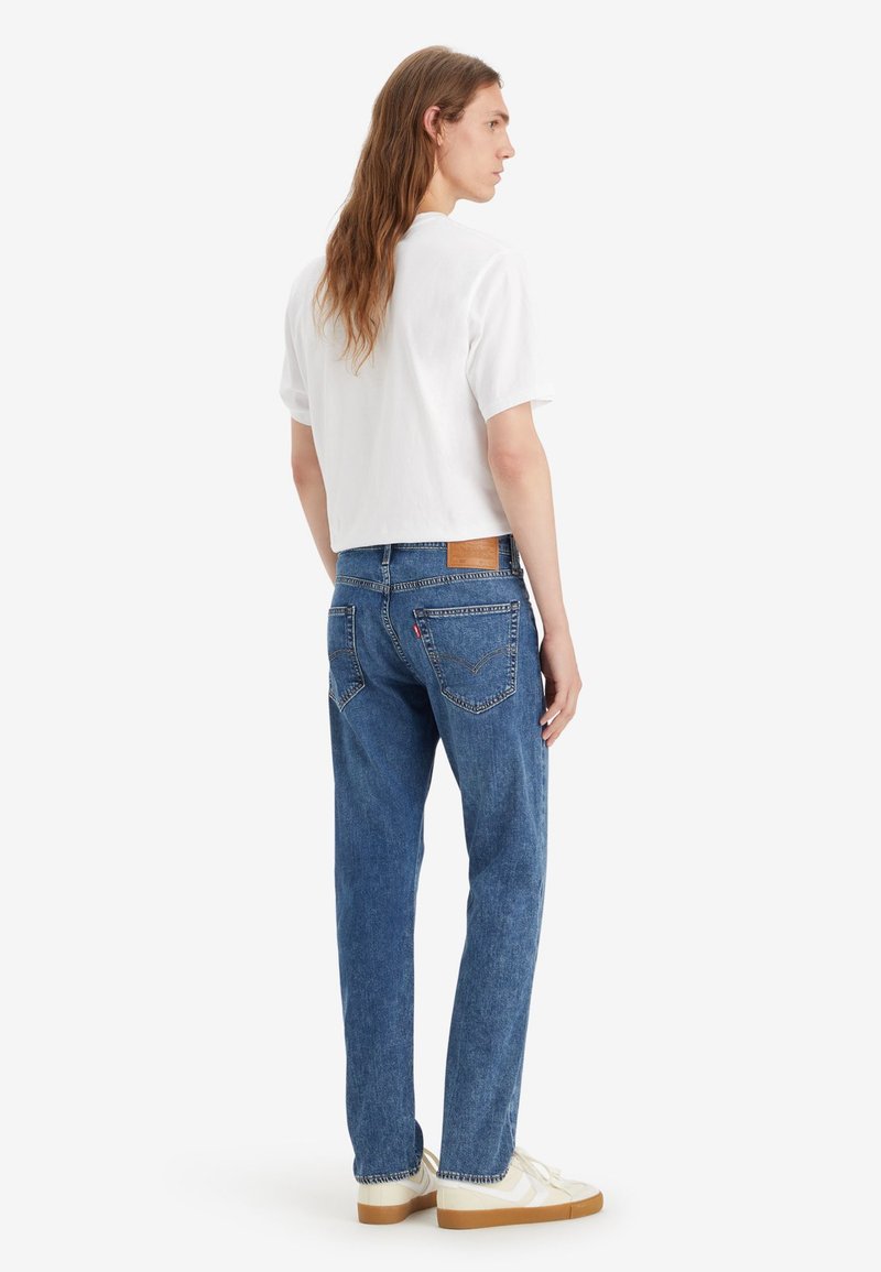 Weißes, kurzärmeliges, cropped T-Shirt, kombiniert mit blauen Jeans und einem Lederpatch am hinteren Bund, dazu weiße Sneakers.