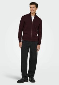 Burgundska pletena zip-up puloverja z visokim ovratnikom, rebrastimi manšetami in kontrastnimi teksturiranimi črtami, v kombinaciji z črnimi ravnimi hlačami in čevlji.