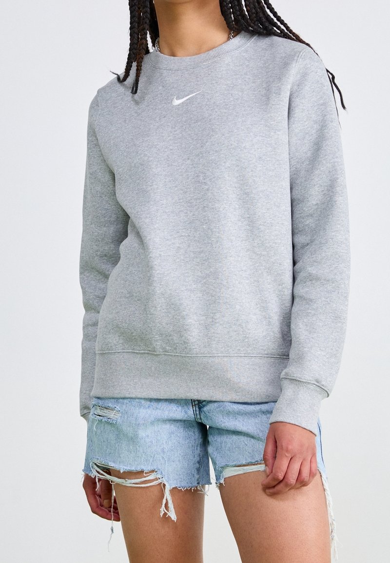 Grå crewneck sweatshirt med ribbade ärmslut och fåll, med en liten vit Nike-logotyp på bröstet. Bärs med slitna denimshorts.