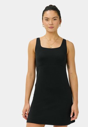 Woman with dark hair in a bun wearing a sleeveless black dress and gold jewelry on a plain light background.