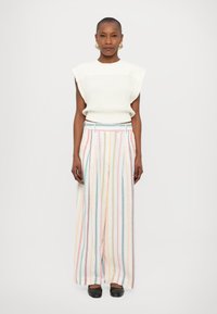 Svetlo pletena brezrokavna majica v off-white barvi, ujemajoča se s široko prugastimi hlačami pastelnih barv, ki imajo pas s pasom in sproščen kroj.